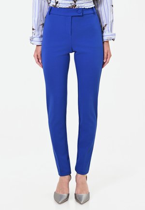 Donna che indossa pantaloni aderenti blu vivace, abbinati a una camicia a maniche lunghe floreale e tacchi argento a punta, in piedi davanti a uno sfondo bianco.