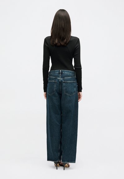 Une femme vue de dos porte un haut noir à manches longues, un jean bleu foncé à jambes larges et des talons hauts noirs à bout ouvert, sur un fond uni.