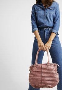 Różowa pikowana torebka na ramię z podwójnymi uchwytami, teksturowaną powierzchnią i metalowymi zdobieniami, trzymana przez osobę w dżinsowym stroju.