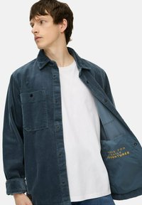 Ung mand iført en blå kordfløjlskappe over en hvid t-shirt, der viser jakkeens inderside med teksten "Lavede til daglige eventyr."
