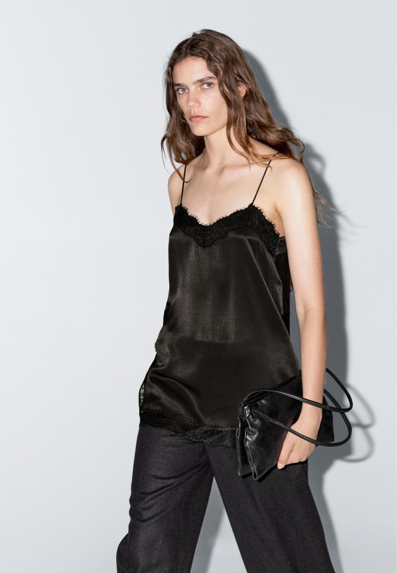 Black silk camisole with lace trim, featuring thin straps, paired with dark gray trousers, and a black handbag. Smooth texture, elegant design.
