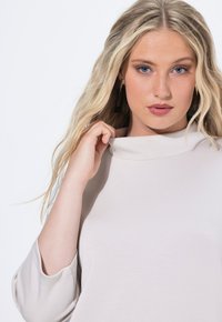 Top beige chiaro con un collo alto e maniche a 3/4, realizzato in un tessuto liscio. Presenta una vestibilità comoda e dettagli di cucitura minimali.