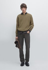 Olijfgroene gebreide trui, zwarte overhemd eronder, grijze jeans, houdt een donkerbruin leren jack vast, zwarte schoenen, minimalistische achtergrond.