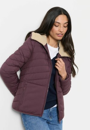 Kvinde med langt mørkt hår iført en lilla quiltet jakke med beige fleecekrave, som lynlåser den op over en hvid skjorte og blå jeans.