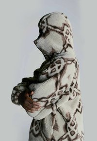 Løs beige hættejakke med brune geometriske mønstre. Har en afslappet pasform, store lommer og tekstureret stof for varme.