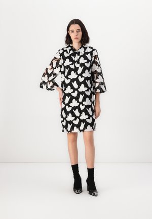 Robe noire et blanche avec des motifs de papillons, manches transparentes, fermeture à boutons et coupe droite. Portée avec des chaussures à talons hauts noires.