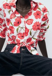 Personne à la peau foncée portant une chemise blanche boutonnée avec des motifs floraux rouges nouée à la taille et un jean bleu foncé.