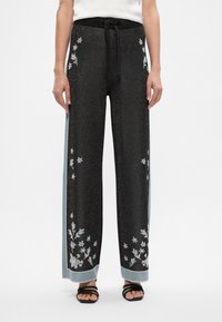 Pantalons noirs à jambes larges présentant une texture scintillante, des broderies florales bleues et des accents bleu clair sur les côtés. Ceinture élastique avec un lacet.