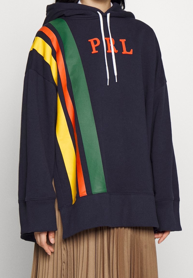 Sweat à capuche marine avec des rayures diagonales jaunes, orange, noires et vertes, et "PRL" en lettres orange sur le devant, porté avec une jupe plissée beige.