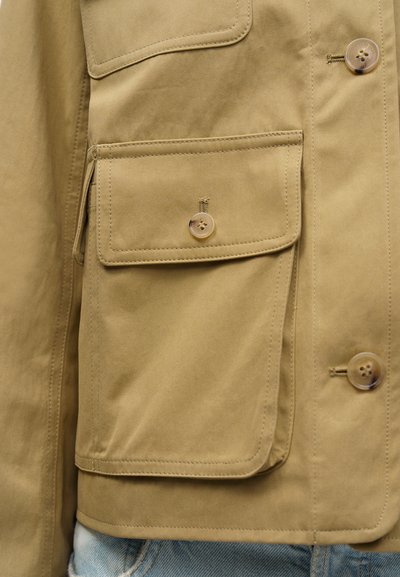 Καφέ τζάκετ με τσέπη με καπάκι και κουμπί, με εμφανείς ραφές, σε συνδυασμό με ανοιχτόχρωμο τζιν παντελόνι.