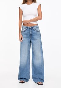Jean en denim bleu clair à jambes larges avec une finition délavée, doté d'une taille haute, de poches avant et d'une fermeture classique à bouton et zip.