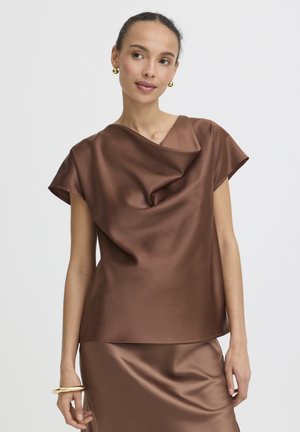 Nő fényes barna szatén ujjatlan felsőben és hozzá illő szoknyában, arany fülbevalóval és karkötővel kiegészítve, egyszínű háttér előtt pózol.