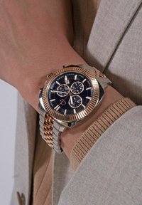 Hronogrāfa pulkstenis ar melnu ciparnīcu, rozā zelta akcentiem un sudraba un rozā zelta nerūsējošā tērauda siksniņu; iekļauj trīs papildu ciparnīcas un datuma rādītāju.