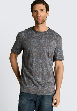 Mężczyzna w ciemnoszarej, wzorzystej koszulce z krótkim rękawem i niebieskich dżinsach, stojący z jedną ręką za plecami i o neutralnym wyrazie twarzy.