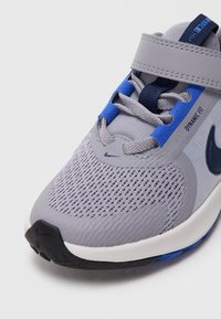 Grijze sportieve schoen met een mesh bovenwerk, verstelbare band, blauwe accenten en een geprofileerde zool voor grip. Nike-logo zichtbaar aan de zijkant.