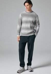 Grauer Gradient-Strickpullover mit gerippten Bündchen und Saum, kombiniert mit dunklen Hosen und grauen Sneakers mit weißen Akzenten und Schnürsenkeln.