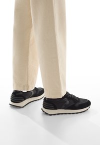 Scarpe sportive nere con materiali testurizzati e lisci, caratterizzate da una suola bianca e sottili accenti a righe sul tallone. Abbinate a pantaloni di colori chiari.