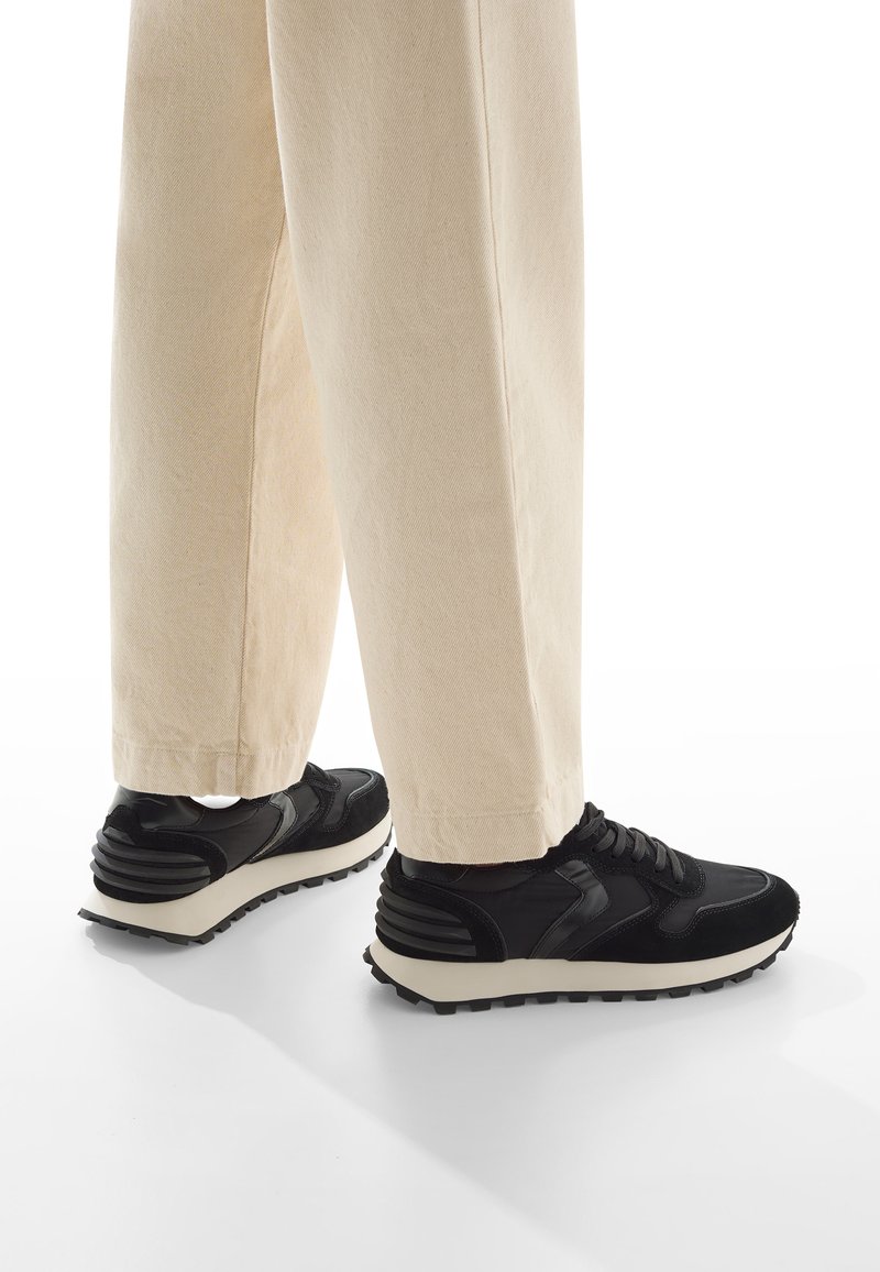 Scarpe sportive nere con materiali testurizzati e lisci, caratterizzate da una suola bianca e sottili accenti a righe sul tallone. Abbinate a pantaloni di colori chiari.