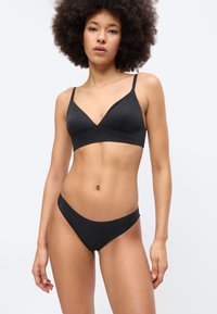 Femme aux cheveux bouclés portant un bralette noir et un slip bikini noir assorti, debout devant un fond clair uni.