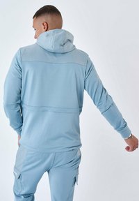 Ljusblå luvtröja med långa ärmar, slät textur, kontrastsömmar och en detalj med söm bak. I kombination med matchande byxor.