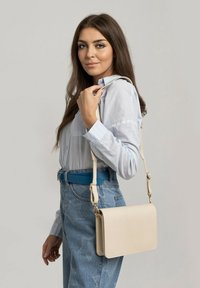 Borsa a spalla rettangolare beige con superficie texture e hardware dorato, portata su una spalla, abbinata a una camicia azzurro chiaro e una cintura blu.