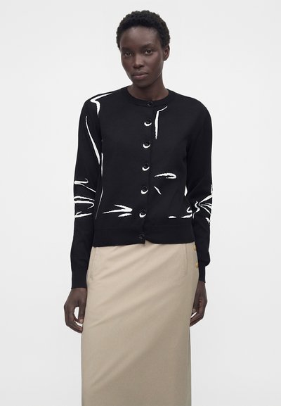 Μαύρη ζακέτα με κουμπιά και άσπρες αφηρημένες γραμμές, φορεμένη με μπεζ φούστα, μοντέλο που στέκεται σε απλό λευκό φόντο.