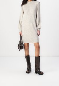 Robe tricotée gris clair avec manches bouffantes, associée à des bottes en cuir brun foncé montant jusqu'aux genoux avec des boucles et un sac à main marron.