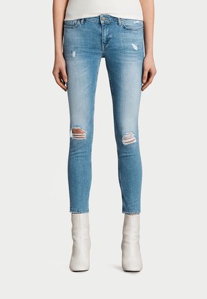 Slim fit -malliset vaaleansiniset farkut polvissa repaleilla, valkoisilla nilkkureilla ja valkoisella yläosalla, kuvattuna keskivartalosta jalkoihin.