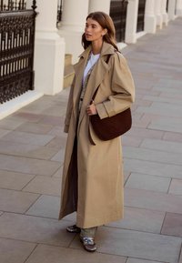 Beige trenchcoat med brede ærmer, lagdelt over en hvid skjorte og mønstrede bukser. Vævet brun skuldertaske og skinnende loafers fuldender looket.