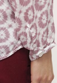 Blusa a maniche lunghe di colore rosa chiaro con motivo tie-dye bianco. Presenta un polsino con bottone bianco e una trama di tessuto liscia e leggera.