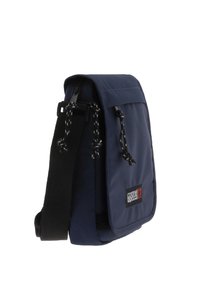 Bolso bandolera azul marino con una solapa, correa negra y acentos decorativos de cuerda. Presenta un parche con el logo en el frente. Fabricado en tela resistente.
