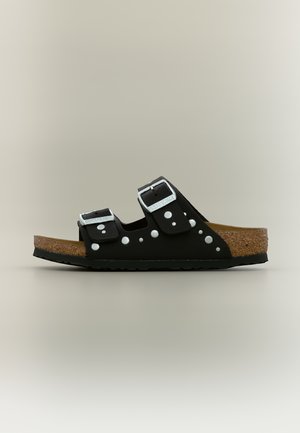 Zwarte open teen sandaal met kurken zool, twee verstelbare zilveren gespen en een wit polkadotpatroon op de bandjes, gezien vanaf de zijkant.