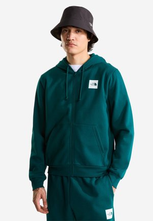 Junger Mann mit dunkelgrünem Zip-Hoodie und passenden Hosen mit weißen Logo-Patches sowie einem schwarzen Bucket Hat.