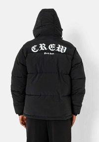 Zwarte pufferjack met een capuchon, voorzien van grote witte tekst "CREW" en "Sixty June" op de bovenrug, gestikte textuur en een losse pasvorm.