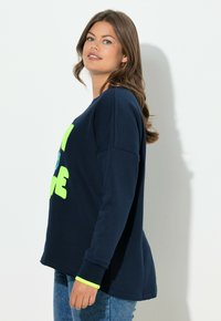 Frau mit langen braunen Haaren, die einen marineblauen Pullover mit neongrüner Schrift und blaue Jeans trägt, seitlich an einer weißen Wand stehend.