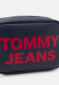 Borsa cosmetica blu navy con lettering rosso "TOMMY JEANS", design rettangolare, texture liscia e zip argentata in cima.