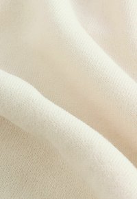 Lichtbeige gebreide stof met een zachte textuur, voorzien van een fijn geweven patroon en zachte vouwen, wat een gladde, subtiele uitstraling creëert.