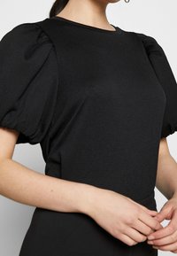 Top nero con maniche a sbuffo, tessuto testurizzato e collo rotondo classico. Il materiale sembra leggero e leggermente rigato.