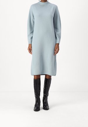 Femme portant une robe midi en maille bleu clair à manches longues, associée à des bottes noires montantes, debout sur un fond blanc.