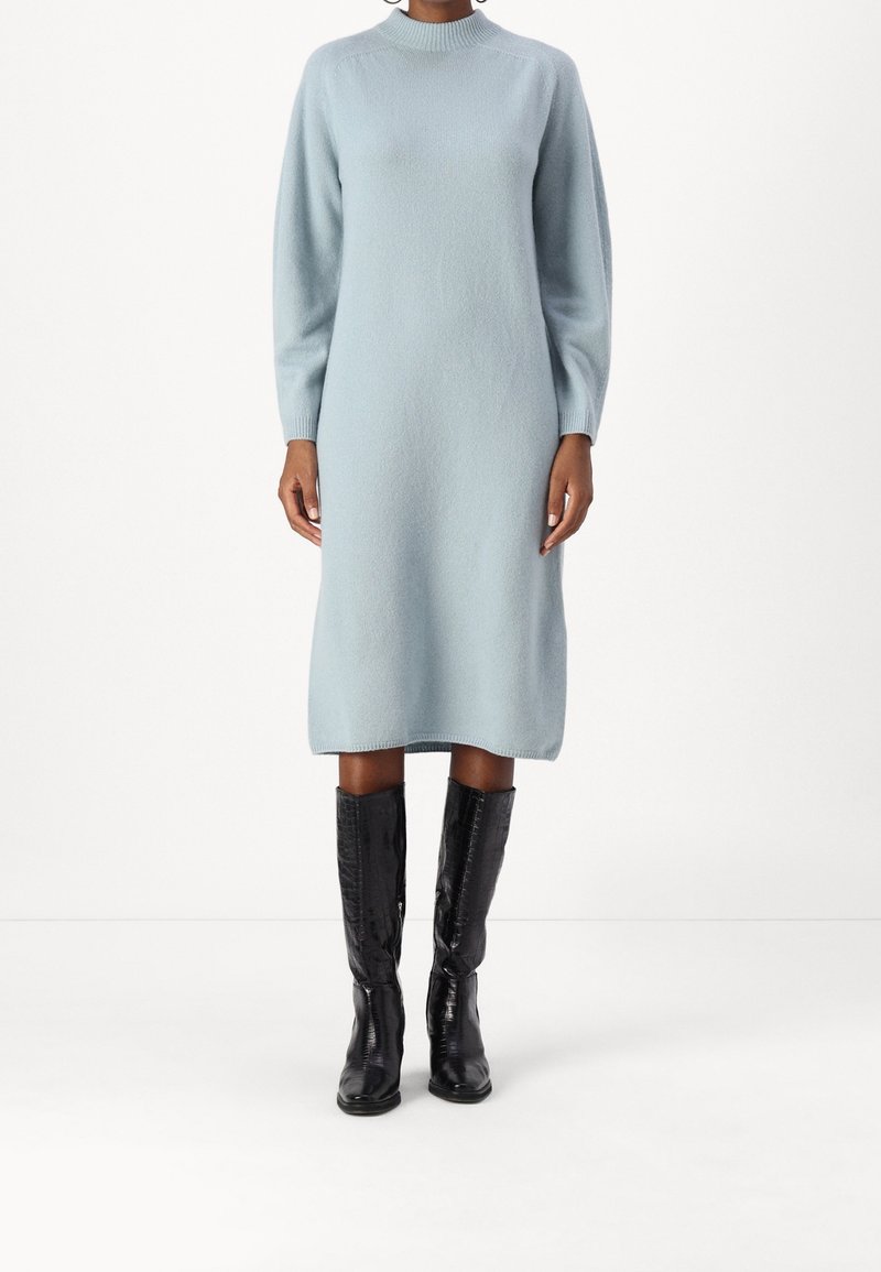 Femme portant une robe midi en maille bleu clair à manches longues, associée à des bottes noires montantes, debout sur un fond blanc.
