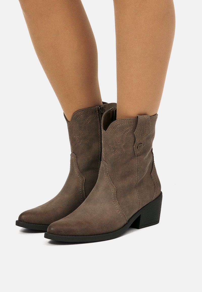 Botines de ante marrón con tacones bajos, costuras decorativas y cremalleras laterales, usados sobre piernas al aire en un fondo sencillo.