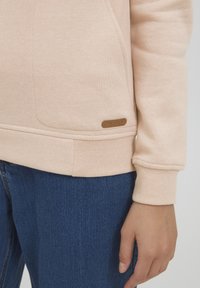 Sweatshirt rosa chiaro realizzato in tessuto morbido con polsini e orlo a costine, caratterizzato da un piccolo tag logo marrone vicino alla parte inferiore sinistra.