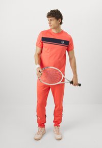 Koral atletik sæt med marineblå vandret stribe, inklusive en t-shirt og matchende joggers. Holder en tennisracket og har lyse sneakers på.