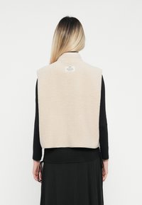 Žena s dlhými blond vlasmi, otočená chrbtom, oblečená v béžovej bezrukávovej fleecovej veste cez čierny top s dlhými rukávmi a sukňu.