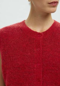 Gilet tricoté rouge avec un col rond, présentant une texture douce et duveteuse. Six boutons à l'avant sont également rouges, complétant l'ensemble du design.