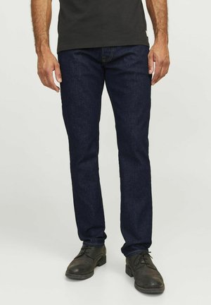Temno modri jeans s slim fit krojem, s klasičnim pet-žepnim dizajnom in šivanim detajlom, v kombinaciji s črnimi elegančnimi čevlji.