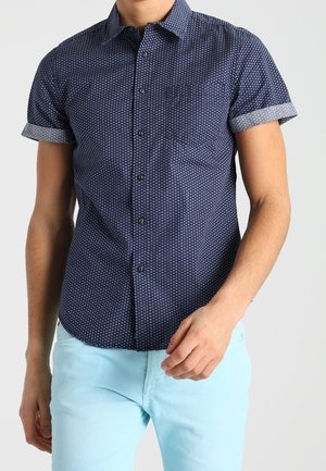 Mężczyzna w dopasowanej, granatowej koszuli na guziki z krótkim rękawem i wzorem, z podwiniętymi mankietami oraz jasnoniebieskich spodniach.