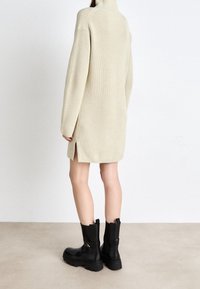 Abito maglione beige con colletto alto, maniche lunghe, spacco laterale e stivali neri alla caviglia con suola spessa e cerniera laterale.