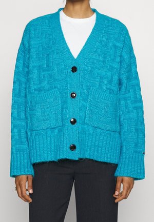 Frau trägt eine hellblaue, strukturierte Strickjacke mit vier schwarzen Knöpfen und aufgesetzten Taschen über einem weißen Hemd und dunkler Hose.