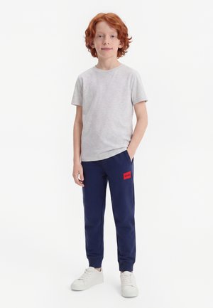 Garçon roux debout portant un t-shirt gris clair, un pantalon bleu marine avec une étiquette rouge sur la cuisse gauche et des baskets blanches.
